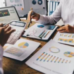 Zwei Personen analysieren Finanzdiagramme und Berichte am Tisch mit Tablet und Taschenrechner während ein Asset-Manager Investitionsdaten auswertet.