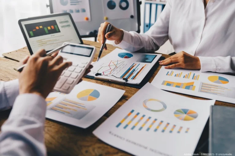 Zwei Personen analysieren Finanzdiagramme und Berichte am Tisch mit Tablet und Taschenrechner während ein Asset-Manager Investitionsdaten auswertet.